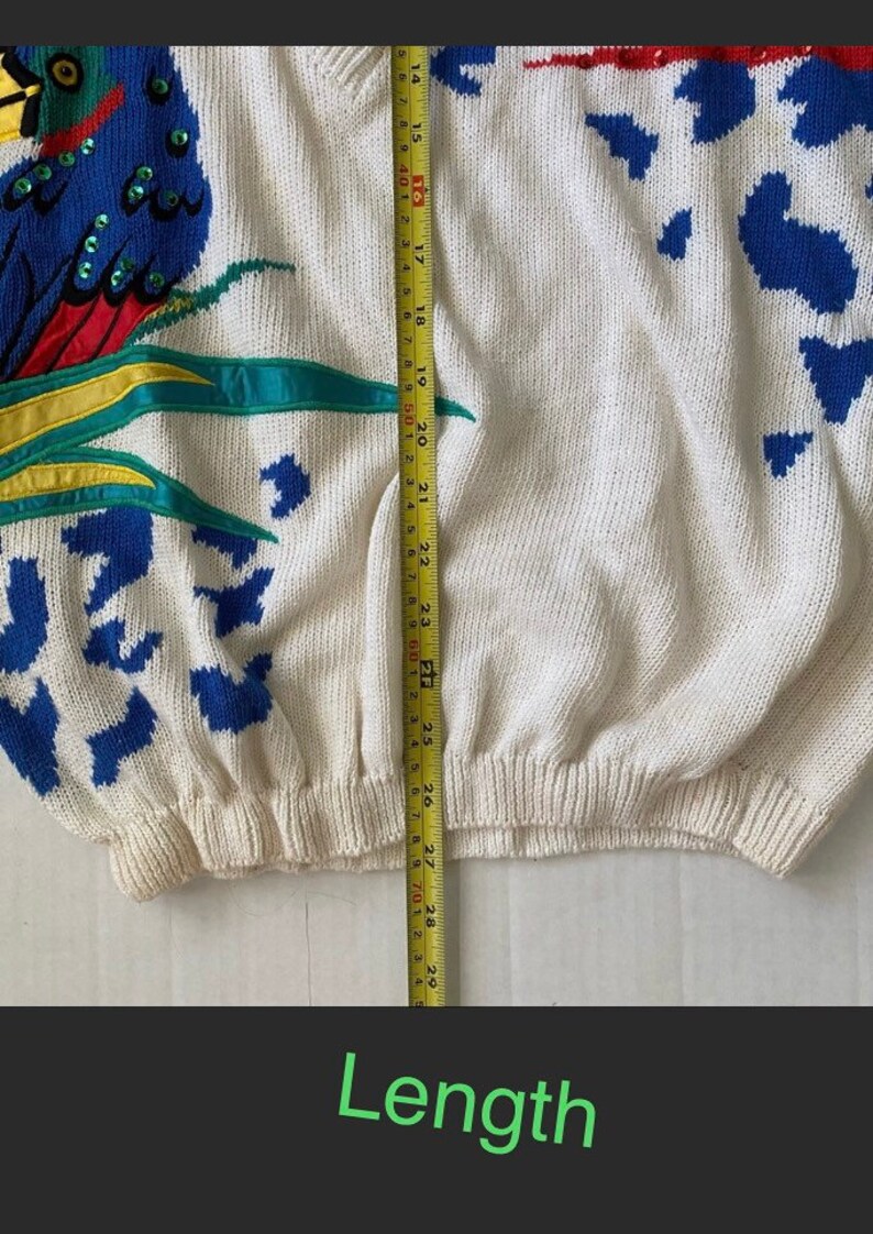 Op de afbeelding: Een witte gebreide trui met een kleurrijk papegaai-ontwerp. De trui heeft een blauwe, rode en gele papegaai met groene bladeren. De trui is 28 inch lang.