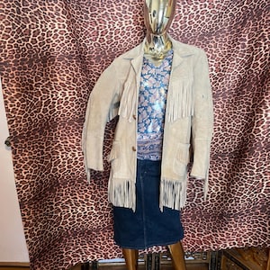 May include: A beige suede jacket featuring long fringe detailing on the chest, sleeves, and pockets, worn over a blue and white floral patterned top and a dark blue denim skirt on a gold-toned mannequin.
