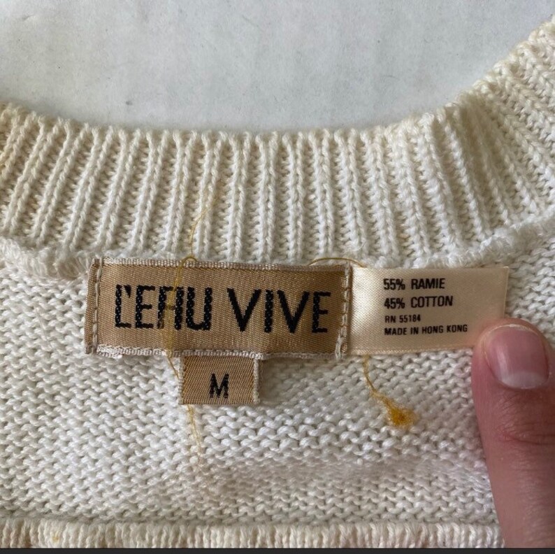Op de afbeelding: Een witte trui met een bruin label waarop "LEAU VIVE" en "M" staat. Het label bevat ook de stofsamenstelling: 55% Ramie en 45% Katoen, RN 55184, en "Gemaakt in Hong Kong".