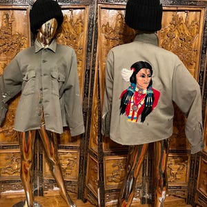 May include: A gray button-up jacket with two chest pockets and paint splatters. The back of the jacket features an embroidered portrait of a Native American woman in red, black, and white. The jacket is displayed on a mannequin.