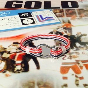 May include: A nautical-style bracelet with red, white, and navy blue rope, secured by a silver-colored clasp. The bracelet is displayed on a reflective surface, with a hockey-themed background.