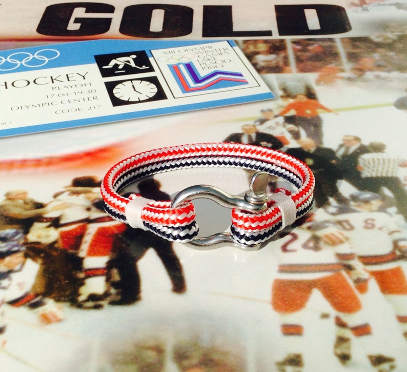 May include: A nautical-style bracelet with red, white, and navy blue rope, secured by a silver-colored clasp. The bracelet is displayed on a reflective surface, with a hockey-themed background.