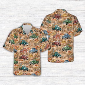 May include: A short-sleeve button-down shirt featuring an all-over print of vintage trucks, red barns, hay bales, and orange pumpkins. The pattern is set against a light tan background, showcasing various farm-themed illustrations in autumn colors.