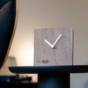 Op de afbeelding: Een vierkante houten klok met een lichte houtnerf afwerking. De klok heeft witte wijzers en een goudkleurig midden. De tekst "WOHOOD" staat op de wijzerplaat. De klok staat op een zwarte tafel.