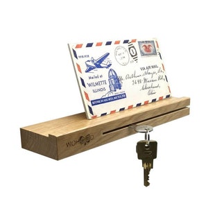 Op de afbeelding: Een houten sleutelhanger met een gleuf voor post. De sleutelhanger heeft een donkere houtkleur en de tekst "WOHOOD" is gegraveerd op de voorkant. Een vintage ansichtkaart met een vliegtuig en de tekst "Mailed at WILMETTE ILLINOIS" wordt in de gleuf weergegeven. Een set sleutels hangt aan een haak aan de zijkant van de sleutelhanger.