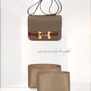 Puede incluir: Un bolso de mano de cuero marrón con una hebilla dorada en forma de H y una correa de cadena dorada. El bolso está sobre una superficie blanca junto a dos organizadores de bolsos de fieltro marrón.