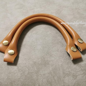 May include: A pair of light brown leather bag handles with brass-colored rivets. The handles are curved and appear to be new, with a smooth texture. The handles are designed for use in crafting or repairing bags.