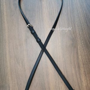 May include: Black crossbody bag strap with silver-tone metal clasps and buckle. The strap is made of a smooth, faux leather material. The strap is shown against a wooden background.