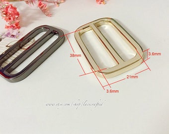 Könnte beinhalten: Zwei Metall-Schieber-Schnallen, eine goldene und eine schwarze, mit Maßen in Zentimetern. Die goldene Schnalle ist 9,1 cm lang, 5,3 cm breit und 0,35 cm dick. Die schwarze Schnalle ist 3,8 cm lang, 2 cm breit und 0,35 cm dick.