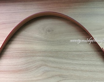 May include: A curved, brown leather strap with a lighter brown interior. The strap has a reddish-brown edge and is attached to the ends with small, light-colored leather loops and metal hardware. The strap is shown on a light wood surface.