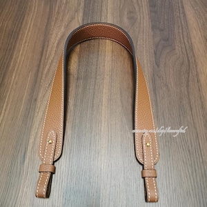 May include: A brown leather bag strap with white stitching and gold-colored hardware. The strap has a textured surface and a curved design, with a loop at each end for attachment.