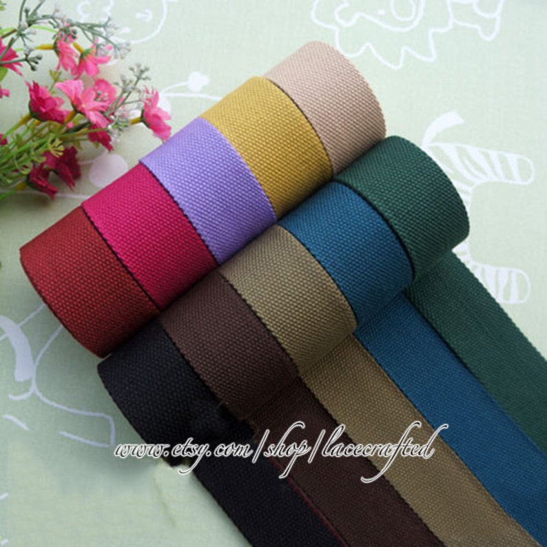 May include: A collection of 10 rolls of cotton webbing in various colors, including brown, black, beige, teal, green, purple, red, and yellow. The webbing is rolled up and stacked on top of each other. The text "www.etsy.com/shop/lacecrafted" is visible at the bottom of the image.