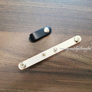 以下が含まれることがあります： 黒と薄いピンクの革製スナップストラップが2つ。シルバー色のスナップが付いています。薄いピンクのストラップは長く長方形で、黒いストラップは短く湾曲しています。濃い木製の表面に置かれています。