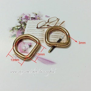 Puede incluir: Dos anillos en D de metal dorado, uno con un diámetro de 13 mm y el otro con un ancho de 3 mm. Los anillos se muestran sobre una superficie blanca con un patrón floral.