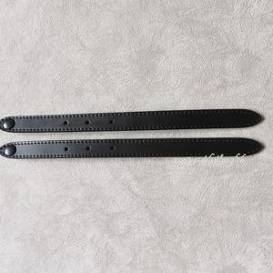 May include: Two black leather straps with button closures. Each strap has a series of small holes and is stitched along the edges. The straps are designed for a bag or other accessory.