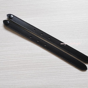 May include: Two long, narrow black leather straps featuring matching black snap buttons at one end. Each strap has a series of evenly spaced holes along its length, with visible stitching running along the edges of the material for a finished look.
