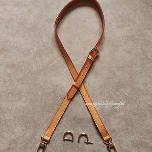 Può includere: Una tracolla per borsa in pelle marrone chiaro con finiture dorate, tra cui due anelli a D e due chiusure. La tracolla è incrociata e ha una fibbia per la regolazione. Lo sfondo è grigio neutro.