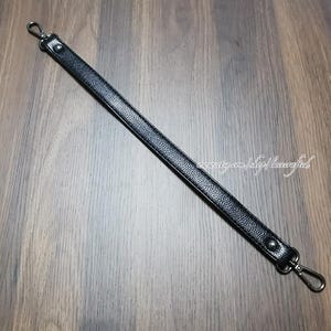 May include: Black leather bag strap with silver-tone metal swivel clasps at each end. The strap is textured and has a rectangular shape. The strap is shown on a wooden surface.
