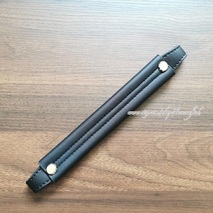 May include: A black leather handle with gold-coloured rivets. The handle has a stitched design down the centre and is shown on a wooden surface. The handle is approximately 25 cm long.