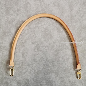 May include: A curved, tan leather bag handle with gold-colored metal clasps. The handle has a rounded shape and is stitched along the edges. The interior edge is a reddish-brown color. The background is a textured gray surface.