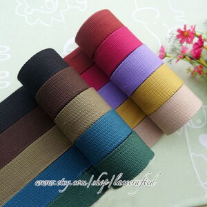 May include: A collection of 10 rolls of cotton webbing in various colors, including black, brown, beige, green, blue, purple, red, and orange. The webbing is rolled up and stacked on top of each other. The text "www.etsy.com/shop/lacecrafted" is visible at the bottom of the image.