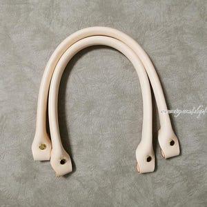 Puede incluir: Dos asas de bolso de cuero color crema con remaches dorados. Las asas son curvas y están unidas en la base. El fondo es gris neutro.