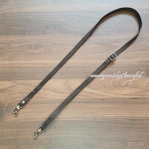 May include: A dark gray adjustable strap with metal swivel clasps and a metal buckle. The strap is made of a woven material and is designed to be attached to a bag or purse. The strap is lying on a wooden surface.