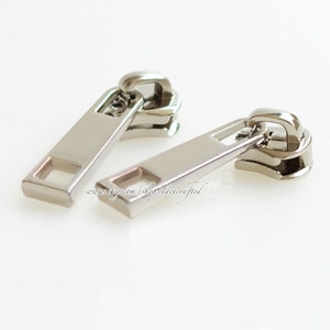 Op de afbeelding: Twee zilveren ritsen met een rechthoekige vorm en een vierkante opening in het midden. De ritsen zijn ontworpen voor gebruik met een rits.