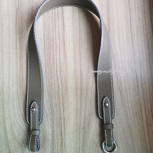 以下が含まれることがあります： 銀色のリングとステッチのディテールが付いた茶色の革のストラップ。ストラップは、バッグの交換用ショルダーストラップとして使用するために設計されています。