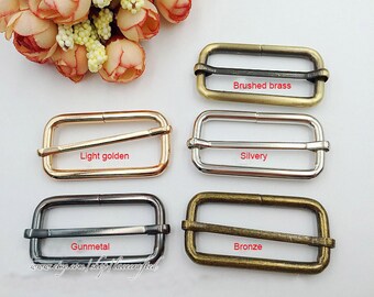 May include: Four metal slider buckles in different finishes: brushed brass, light golden, gunmetal, and bronze. Each buckle has a rectangular shape with a center bar.
