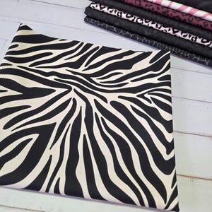 Könnte beinhalten: Ein gefalteter Stoff mit einem auffälligen schwarz-weißen Zebrastreifenmuster. Im Hintergrund ist ein Stapel verschiedener Stoffe mit Tiermustern in Schwarz, Rosa und Weiß zu sehen. Geeignet für Näharbeiten und Bastelprojekte.