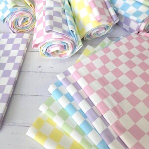 Puede incluir: Una colección de rollos de tela y piezas plegadas con un patrón de cuadros repetitivo en colores pastel, incluyendo rosa claro, lavanda, amarillo pálido y azul bebé, dispuestos en una pila y paquetes enrollados sobre una superficie blanca.