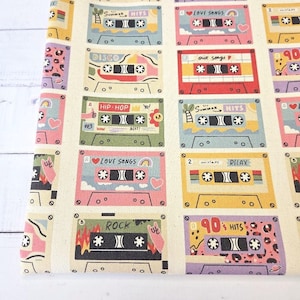 Könnte beinhalten: Ein cremefarbener Stoff mit einem sich wiederholenden Muster aus bunten Musikkassetten. Jede Kassette zeigt einzigartige Designs und Textbeschriftungen wie Summer Hits, Love Songs, Disco, Hip-Hop, Relax, Rock und 90s Hits in verschiedenen Farben.
