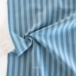 Könnte beinhalten: Ein Stück Webstoff mit einem sich wiederholenden vertikalen Streifenmuster in abwechselnden Schattierungen von Hellblau und Mittelblau. Das Textil weist eine sichtbare Webstruktur auf und enthält das Wort FABLEISM in kleinen weißen Buchstaben.