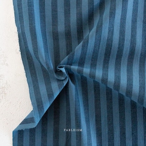 Könnte beinhalten: Ein Stoffstück mit einem vertikalen Streifenmuster in abwechselnden Schattierungen von dunkelmarineblau und hellerem Himmelblau. Das Textil hat eine sichtbare Webstruktur und enthält das Wort FABLEISM, das in kleinen weißen Buchstaben unten aufgedruckt ist.
