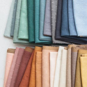 Pode incluir: Uma coleção de amostras de tecido dobradas dispostas em duas fileiras. A fileira superior apresenta tons frios, incluindo azul claro, verde-azulado, cinza e azul marinho. A fileira inferior exibe tons quentes como rosa, terracota, laranja, creme e amarelo. Cada peça tem um pequeno padrão de ponto cruz branco.