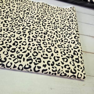 Puede incluir: Tela doblada con un fondo color crema y un patrón repetido de manchas de leopardo negras. La tela es probablemente de algodón o un material similar, adecuada para proyectos de costura o decoración del hogar.