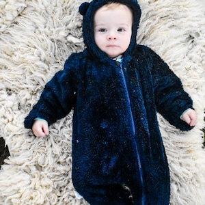 May include: A dark navy blue fuzzy hooded jumpsuit with long sleeves and integrated feet. The garment features a central zipper closure running from the neck down to the torso, and the hood includes small rounded ears on the top corners.