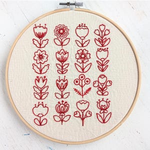 May include: A hand embroidery kit titled FOLK FLORALS REDWORK featuring a circular wooden hoop holding off-white fabric with red stitched floral patterns. The kit includes pattern transfer paper, thread, fabric, instructions, and video links.
