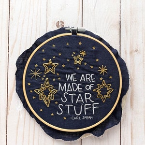 May include: Embroidered hoop art with a navy blue background and gold stars. The text reads "We are made of star stuff - Carl Sagan".