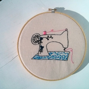 Può includere: Disegno di una macchina da cucire ricamata su un tessuto beige teso su un telaio da ricamo in legno. La macchina da cucire è nera con dettagli blu e bianchi. Il filo è rosa.