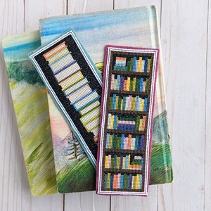 May include: Two handmade bookmarks featuring a bookshelf design. One bookmark is black with white and blue books, the other is pink with colourful books. Both bookmarks are made with cross-stitch.