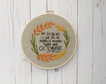 May include: Embroidered hoop art with a beige background and a wreath of green and orange leaves. The text reads "I'm so glad I live in a world where there are Octobers".