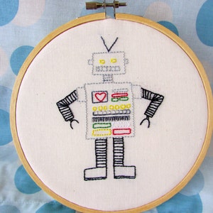 May include: Embroidered hoop art featuring a white robot with black arms and legs, yellow, green, and red details, and a red heart on its chest.