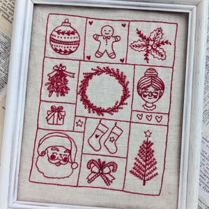 May include: A framed embroidery piece with a white background and red stitching. The embroidery depicts various Christmas symbols, including a Christmas tree, a wreath, a stocking, a gingerbread man, a candy cane, a present, a holly sprig, a mistletoe sprig, a Christmas ornament, and Santa Claus.