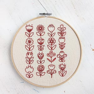May include: A circular wooden embroidery hoop containing off-white fabric with four vertical rows of red thread floral patterns. Each row features distinct, stylized flower and leaf designs stitched in a consistent red outline style.