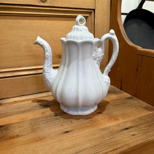 May include: A white ceramic teapot with a lid and a curved spout. The teapot has a ribbed design and a handle. The spout and handle are decorated with floral details. The teapot is sitting on a wooden surface.