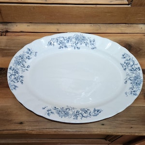 May include: An oval-shaped white ceramic serving platter featuring a scalloped edge and a decorative blue floral pattern printed along the rim. The design consists of clusters of blue flowers and leaves arranged symmetrically around the border.