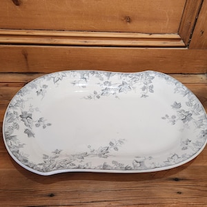 May include: An oval-shaped white ceramic serving platter featuring a decorative gray botanical pattern of leaves and berries along the scalloped edges. The platter has a smooth, glossy finish and a wide, shallow basin for serving food items.