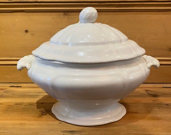 May include: A white ceramic tureen with a matching lid, featuring a rounded body, a pedestal base, and two side handles. The lid is topped with a small, smooth knob, and the entire piece has a glossy finish with subtle, molded panel detailing.
